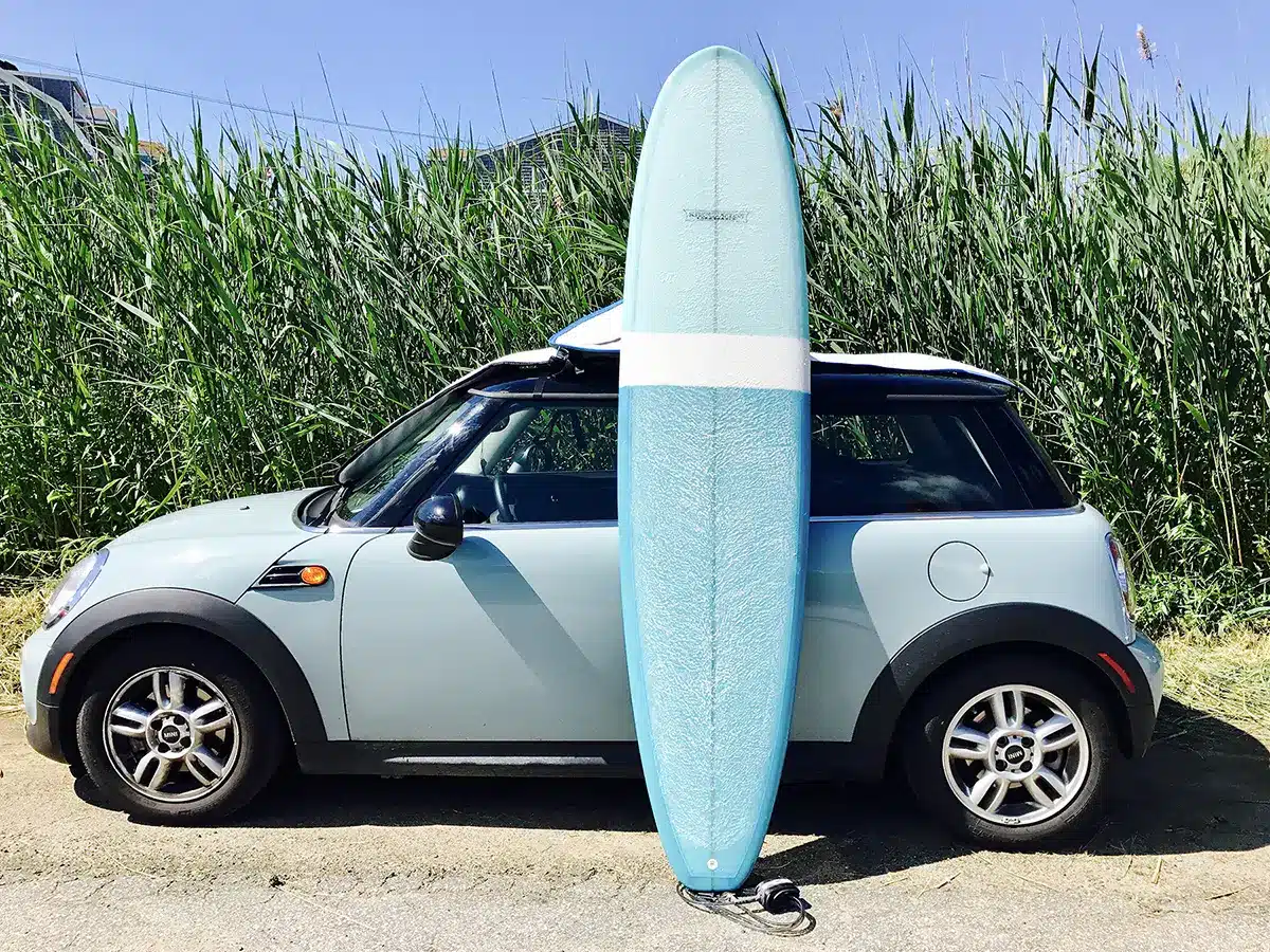 Blue longboard surfboard leaning against a light blue Mini Cooper parked beside tall beach grass